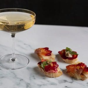 Close-up of slices of beetroot-cured trout on slices of crusty bread, served with a glass of fizzy wine.